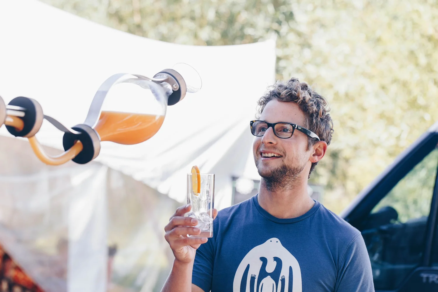 Rik Turner holding a drink outdoors with white tents and trees in the background, smiling